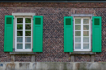 Fototapeta na wymiar Beautiful vintage exterior of building with old rough brick wall facade and elegant windows which consist of wooden green shutters, deluxe white concrete sash and frieze, and small balcony.