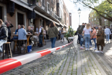 Selected focus view at Red and white caution tape restrict outdoor seating area in old town D&uuml;sseldorf, Germany during lockdown. Crowd of people enjoy reopening restaurant during epidemic COVID-19. 