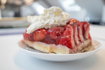 Selective focus, close-up view at traditional German sliced fresh strawberry cake tart or pie on topped with whipped cream, and defocused background of white interior room. 