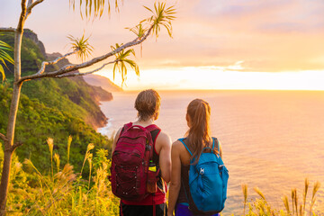 Hawaii hiking hikers hiking on Kalalau trail watching sunset from Na Pali Coast. Tourists couple with backpacks walking outdoor in Kauai island. Summer travel adventure active lifestyle.