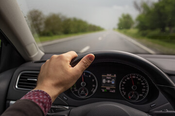 Close-up of male hand holding steering wheel, driving the car. Background of blurred road in rainy day. Travel concept.