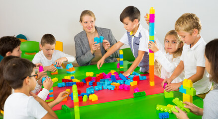 Young female teacher and happy schoolkids building castle from plastic toy blocks during lesson in...