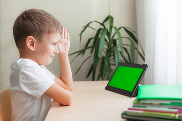 Smiling school boy siting at desk and looks at tablet with green screen. A child pulls a hand to answer. Online lesson at a distance learning. Green screen chroma key on the monitor. Side view