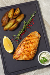 Grilled chicken breast with rosemary and lemon, baked potato wedges and sauce on a dark platter. Summer menu concept. Healthy eating. View from above