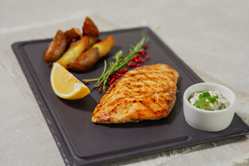 Grilled chicken breast with rosemary and lemon, baked potato wedges and sauce on a dark platter. Summer menu concept. Healthy eating