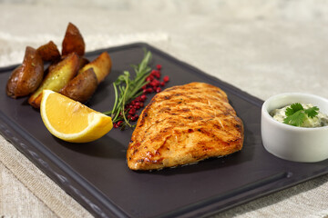 Grilled chicken breast with rosemary and lemon, baked potato wedges and sauce on a dark platter. Summer menu concept. Healthy eating