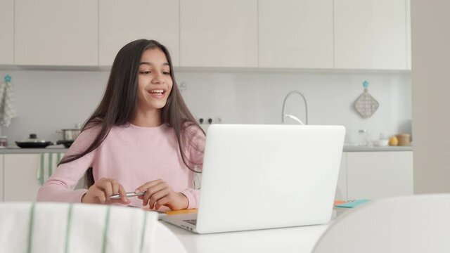 Cute indian latin arab teen girl e learning at home using pc laptop. Smart kid raising hand on remote distant lesson answering teacher's questions writing notes. Remote school education concept.