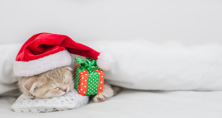 Cute kitten wearing red santa's hat sleeps with gift box under warm white blanlet on a bed at home.Empty space for text