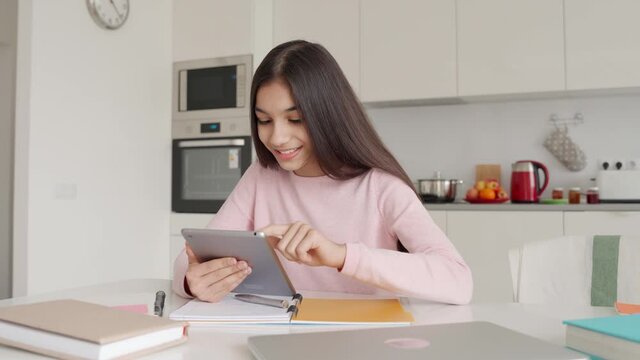 Pretty indian latin arab teenage happy smiling schoolgirl doing online interactive gaming homework e learning at home using computer tablet sitting at kitchen table. Remote school education concept.