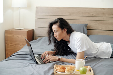 Serious modern young Vietnamese guy in white tshirt working with laptop remotely while lying in bed...