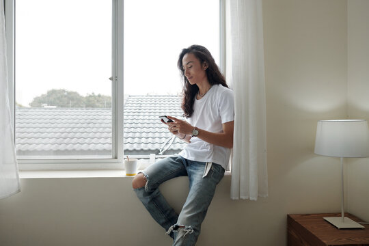 Content young Vietnamese man with wavy hair sitting on window sill and using smartphone while drinking morning coffee in hotel or at home