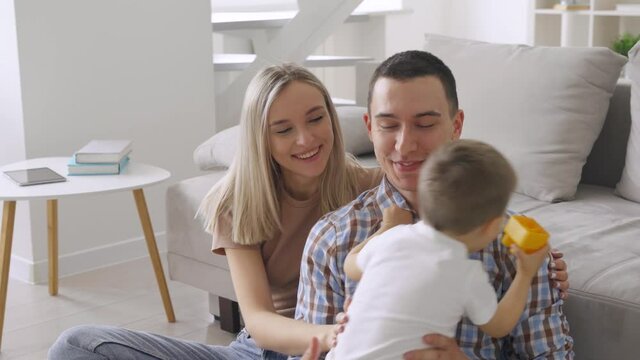 Happy young family with boy child playing enjoying time together at home in living room, kid running to parents' embrace on floor near sofa, mom and dad holding and kissing boy, laughing having fun. 