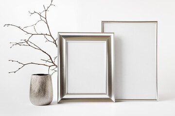 Two silver frames with blank copy space, silver pot, glass vase and dry plant branch.