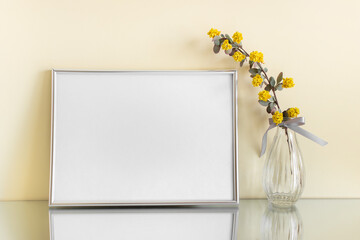 Extra large blank A4 silver frame with yellow summer flower branch in glass vase on mirror surface.