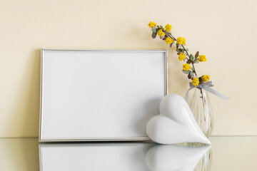 Romantic composition with extra large silver blank frame and white porcelain heart, yellow flowers on mirror reflective surface.