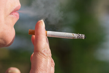 Middle age woman smoking cigarette outdoors with sun on her face. Closeup woman with cigarette in her mouth copy space.