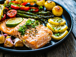 Roasted salmon steak with roast potatoes and vegetable salad served on black plate on wooden table
