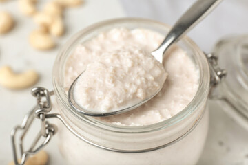 Jar and spoon with cashew sour cream on light background, closeup