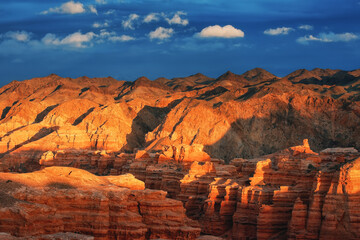 Sunset in the canyon, sunrays on the mountains, erosion, plateau, cloudy sky and mountains on the horizon. Red rocks and layers. Charyn canyon in Kazakhstan