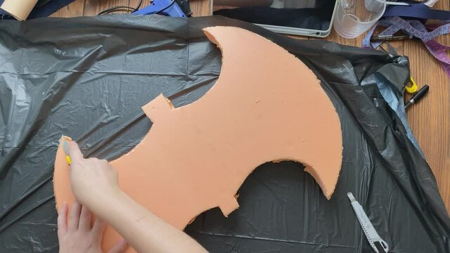 Hands cut axe form from styrofoam with stationery knife on garbage bag. Woman makes viking hatchet for cosplay festival. Diy celtic weapon on wooden table. Craftsman's workshop.