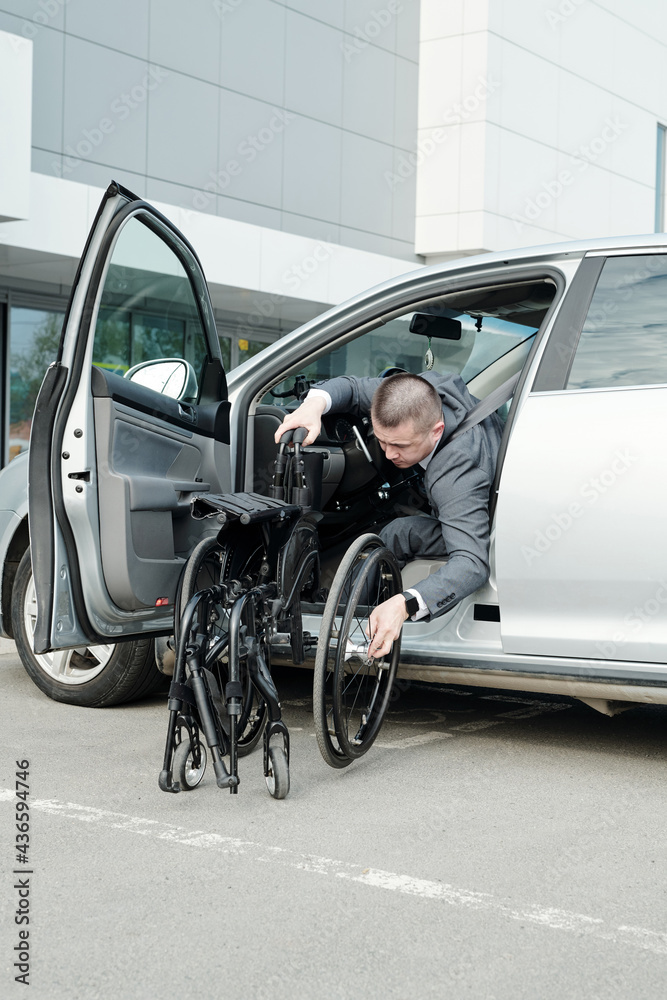 Poster young disabled man in suit taking his wheelchair out of car