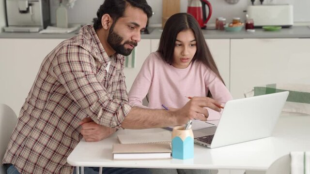 Young indian dad helping teenage daughter schoolgirl study online at home sitting at kitchen table at home using pc laptop computer. Father and kid e learning together having remote distant lesson.