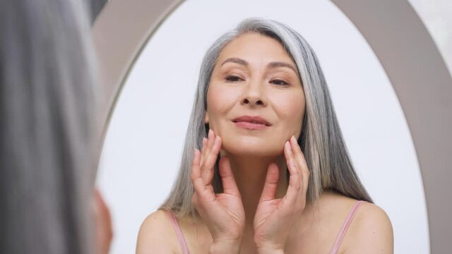 Senior older middle aged Asian woman with grey hair with radiant face with perfect skin looking at mirror smiling to her reflexion touching massaging skin. Ads of senior dry skin care treatment.