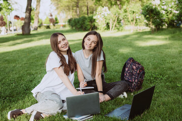 Two female students are sitting in the park on the grass with books and laptops, studying and preparing for exams. Distance education. Soft selective focus.