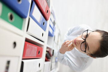 Pensive woman in glasses looks at closet with folder of documents