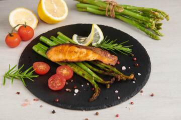 Delicious seasonal green asparagus and sliced smoked salmon on a rustic plate with lemon, salt, pepper, of lemon on a slate. Baked salmon garnished with asparagus and tomatoes with herbs.
