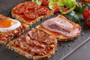 Variety of open sandwiches made of brown whole wheat  bread with tomatoes sauce, tomatoes, white beans, bacon, fried egg and smoked ham. Fresh fruit and vegetable salad
