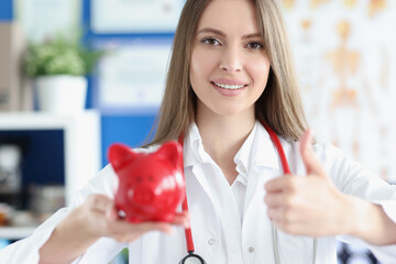 Smiling medical doctor woman holding red pig piggy bank in her hands and gesture thumbs up