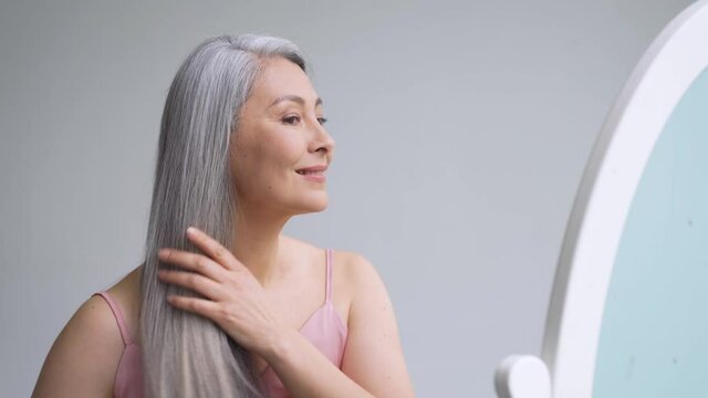 Senior attractive middle 50 years aged asian woman with gray hair looking at mirror reflection combing tangled gray hair. Alopecia hair loss prevention treatment after menopause advertising concept.