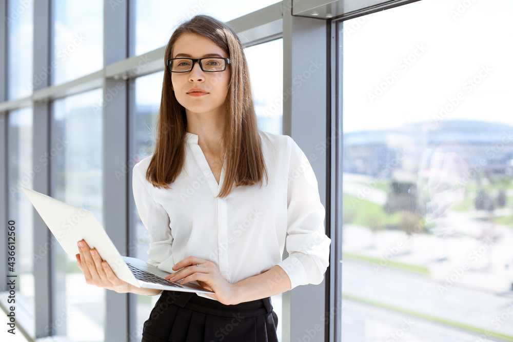 Wall mural beautiful female specialist with laptop computer standing in modern office and smiling charmingly. w