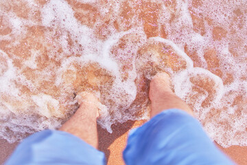Men's feet on beach. Person in jeans shorts stands and looks down at his feet in sand that washes...