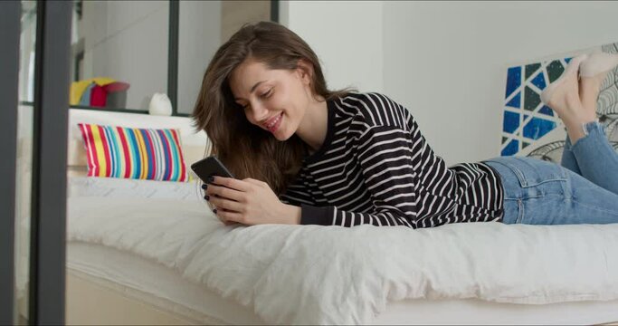 Close-up of attractive caucasian teen girl lying on bed at home and texting on her phone. Tired beautiful young woman is bored at home. 4K RAW edited footage