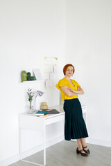 red haired woman in yellow blouse stands near white table against background of white wall and looks at camera. Distant work. Online training. Home office