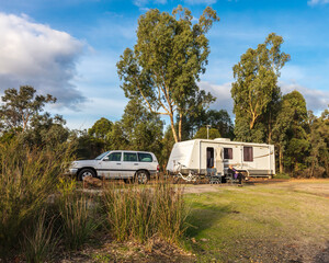 A mature woman sitting outside a large modern caravan and four wheel drive against a background of gum trees under a bright blue sky - obrazy, fototapety, plakaty
