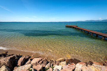 Obraz premium Beautiful beach with a small empty wooden pier on Lake Garda (Lago di Garda) between the small villages of Lazise, Bardolino and Cisano. Tourist resort in Verona province, Veneto, Italy, Europe.