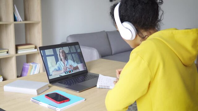 African American mixed race adult student in headset having virtual meeting online call educational webinar with senior teacher at home office writing notes. Video e learning conference call on pc.