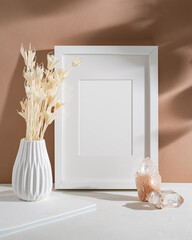 Empty frame with mat, white vase with dried flowers, crystals illuminated by sunlight. Mock up poster with place for your design in the terracotta interior.