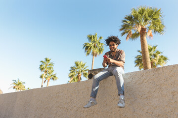 Happy Afro man having fun using mobile smartphone while listening to music with vintage boombox stereo in tropical place during summer vacations