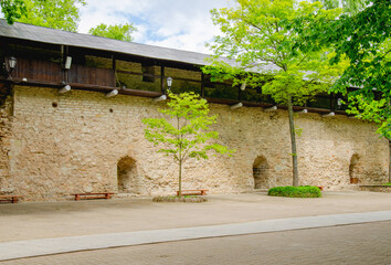 The monastery wall, benches and trees grow against the wall. Clouds are visible above the roof of the wall.