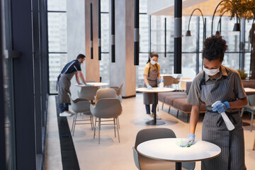 Wide angle shot of workers wearing masks while cleaning tables in cafe interior, covid safety...