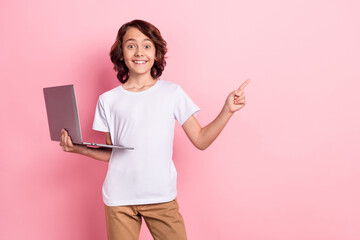 Photo of attractive cheerful small boy point finger empty space hold computer isolated on pink...