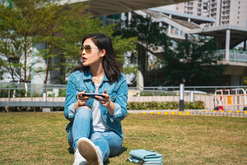 Fototapeta premium Young woman playing smartphone in summer park wearing denim shirt