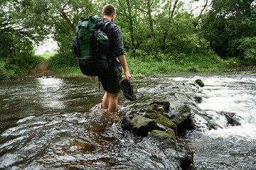 Low section shot of a man in a plaid shirt with heavy hiking backpack crossing the streem barefoot
