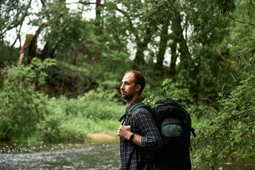The man with a large hiking backpack standing on the bank of the river and enjoying nature