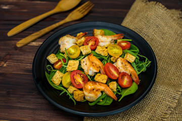 Top view flat lay shot of a homemade grilled shrimp salad with croutons, red and yellow cherry tomatoes in the black dish on wooden table with a wooden spoon and fork. Concept of delicious clean food