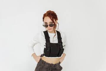 an attractive young red-haired woman wearing sunglasses and a black vest on a white background. bold and daring style in clothing for women casual